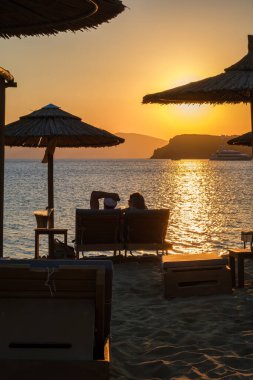 Ios, Yunanistan - 13 Eylül 2022: Güneşli yataklarda uzanan, şarap içen ve Yunanistan 'daki Mylopotas plajında inanılmaz turuncu günbatımının tadını çıkaran bir çiftin görüntüsü