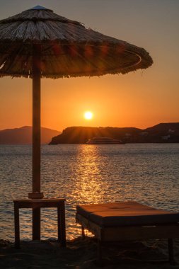 Yunanistan 'da Mylopotas sahilinde güneşli bir yatak, güneş şemsiyesi ve çarpıcı turuncu bir gün batımı.