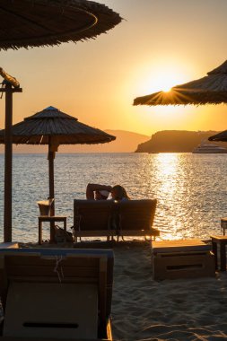 Ios, Yunanistan - 13 Eylül 2022: Güneşli yataklarda uzanan, şarap içen ve Yunanistan 'daki Mylopotas plajında inanılmaz turuncu günbatımının tadını çıkaran bir çiftin görüntüsü