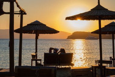 Ios, Yunanistan - 13 Eylül 2022: Güneşli yataklarda uzanan, şarap içen ve Yunanistan 'daki Mylopotas plajında inanılmaz turuncu günbatımının tadını çıkaran bir çiftin görüntüsü