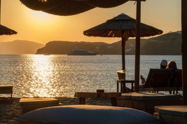 Ios, Yunanistan - 13 Eylül 2022: Güneşli yataklarda uzanan, şarap içen ve Yunanistan 'daki Mylopotas plajında inanılmaz turuncu günbatımının tadını çıkaran bir çiftin görüntüsü