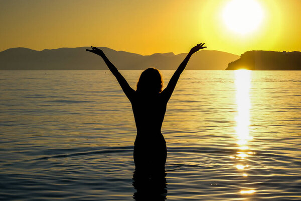 View of a woman standing in the sea  with her hands up in the sky enjoying the amazing sunset at the Mylopotas beach in Ios Greece