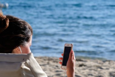 View of a young woman at the beach using her mobile phone and her earphones in Ios Greece
