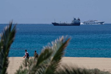 os, Yunanistan: 29 Mayıs 2021: Yunanistan 'daki Mylopotas plajında yürüyen turistleri görmek, arka planda bir kargo gemisi ve yüksek hızlı bir feribot