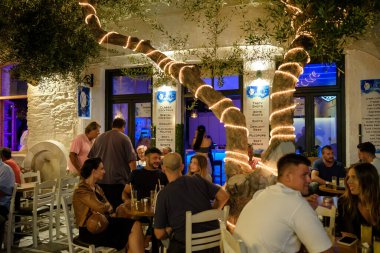 Ios, Yunanistan - 8 Eylül 2023: Barlarla çevrili turistler, gece vakti Yunanistan 'daki popüler meydanda içki ve kokteyllerin keyfini çıkarıyorlar