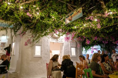Ios, Yunanistan - 9 Eylül 2023: Turistler, Yunanistan 'da renkli sandalyeler ve begonvillerle birlikte açık havada güzel bir terasta içki içiyorlar