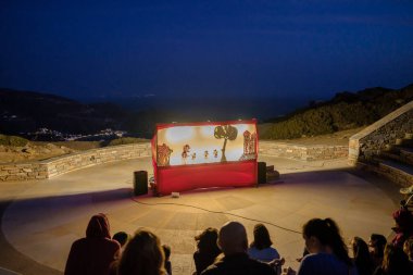 Ios, Yunanistan - 10 Eylül 2023: Odysseas Elytis açık hava tiyatrosunda geleneksel bir kukla gösterisi ve arka planda Yunanistan 'daki ünlü Mylopotas plajı