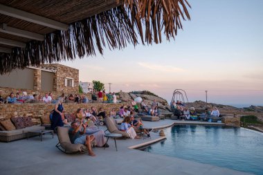 Ios, Yunanistan - 17 Eylül 2023: Ios Yunanistan 'da günbatımının keyfini çıkaran bir salon barında çeşitli insanların yüzme havuzunun önünde oturması