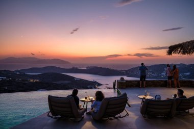 Ios, Yunanistan - 17 Eylül 2023: Yunanistan 'da muhteşem günbatımının tadını çıkaran bir çiftin yüzme havuzunun önünde oturması