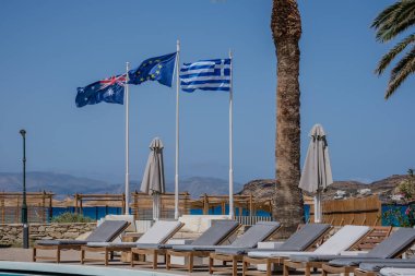 Ios, Yunanistan - 2 Mayıs 2024: Palmiye ağaçları, güneş yatakları ve Ios Yunanistan 'daki ulusal bayraklar ve Avrupa toplumunun yer aldığı güzel bir açık yüzme havuzu manzarası
