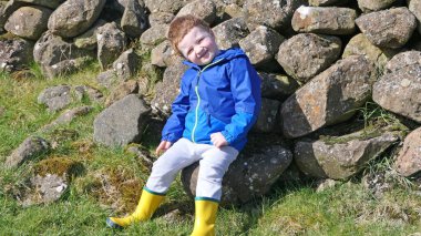 Kırmızı saçlı, mavi ceketli ve sarı çizmeli bir çocuk çiftlikte oynarken eğleniyor. 