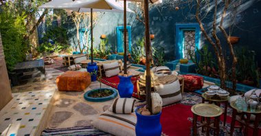 Birçok masası, yastığı ve şemsiyesi olan bir teras. Sahne işlevsel ve davetkar bir açık hava yemek alanını gösteriyor. Oturma seçenekleri ve güneşten uzak gölgeler de var..