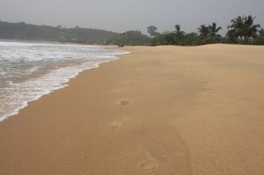 Sahilin kenarından süzülen ayak izlerinin yakın çekimi, yumuşak dalgalar kuma vuruyor. Ayak izleri yalnızlık ve yansıma hissi yaratıyor..