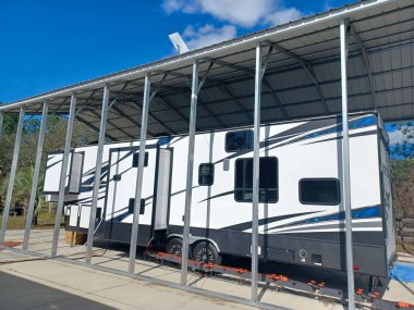 Large RV recreational vehicle under a carport with satellite dish on top.  Temporary extended living setup. Toy hauler style