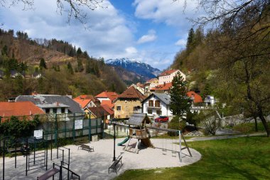 Slovenya 'nın Gorenjska kentindeki Trziç kasabasındaki binaların manzarası. Önünde bir oyun alanı, arkasında karla kaplı bir dağ zirvesi var.