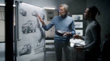 İki farklı otomotiv erkek tasarımcısı ellerinde küçük tekerlekler, fütüristik elektrikli araba tasarımı geliştiriyorlar, çizimlerle birlikte beyaz tahtanın yanında duruyorlar. Modern araba tasarım stüdyosunda çalışıyorlar..