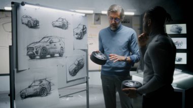 İki farklı otomotiv erkek tasarımcısı ellerinde küçük tekerlekler, fütüristik elektrikli araba tasarımı geliştiriyorlar, çizimlerle birlikte beyaz tahtanın yanında duruyorlar. Modern araba tasarım stüdyosunda çalışıyorlar..