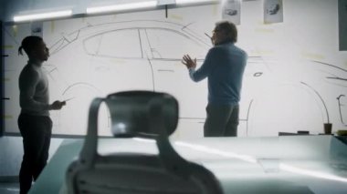 İki farklı otomotiv erkek tasarımcısı gelecekteki elektrikli araba tasarımını tartışıyor ve geliştiriyor. Biri beyaz tahtaya çizimler çiziyor, diğeri tablet bilgisayarı tutuyor. Araba tasarım stüdyosu.
