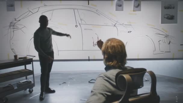 2人の多様な自動車男性デザイナーがスケッチの青写真を議論し ホワイトボード上の未来的な電気自動車の未来のデザインを開発します 彼らは現代の車の設計開発スタジオで働いています — ストック動画