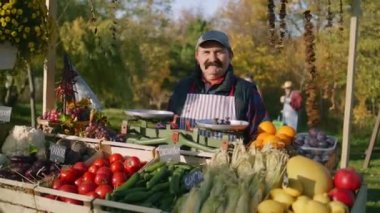 Kıdemli çiftçi elinde taze meyve ve sebzelerle tezgahta duruyor ve kameraya bakıyor. Hafta sonu yerel çiftçi pazarında alışveriş. Vejetaryen, organik ve sağlıklı yiyecekler. Tarım.