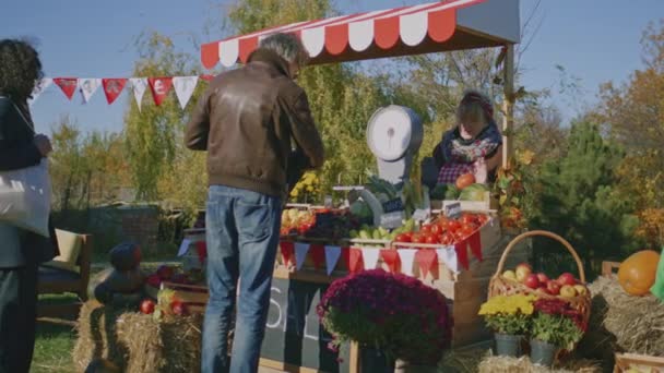 サングラスの成熟した男は新鮮な野菜や果物を選択し それらを量ります セールスポイントのホルダーは 独自の製品を提供しています 週末には地元のファーマーズマーケットで買い物 ベジタリアン オーガニックフード — ストック動画