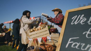 Asyalı kadın ve Afrikalı Amerikalı erkek yerel çiftçi pazarından alışveriş yapıyor. Çift yumurta, meyve ve sebzeleri kağıt eko torbalara doldurur. Hafta sonları açık havada sonbahar festivali. Satış sistemi noktaları.