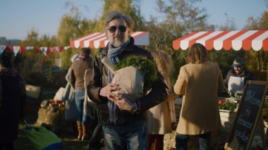 Güneş gözlüklü, elinde meyve ya da sebzeyle poz veren yetişkin bir adam kameraya bakar, pazarda dükkanlar açar, yeni ürünler alır ve sağlığına dikkat eder.