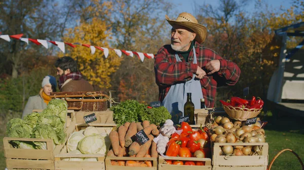 Stock image Senior farmer seller stands at the stall with fruits and vegetables, counts cash money. People shopping at local farmers market. Autumn fair. Organic food. Agriculture. Points of sale system.