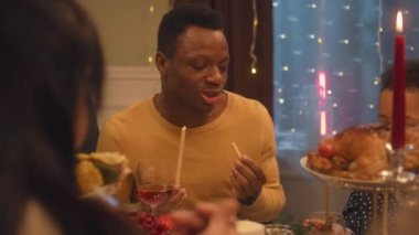 Noel 'i ya da 2023 yılını kutlayan çok kültürlü bir aile. Afro-Amerikalı adam konuşur ve yer. Bulaşık ve mumlarla tatil masası servis edildi. Aile içinde sıcak bir Noel yemeği..