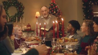 Drandfather aile Noel yemeğinde İncil okur. Noel 'i ya da 2023 yılını kutlamadan önce dua eden mutlu büyük aile. Lezzetli yemek ve mumlarla servis edilen tatil masası. Kış tatili.