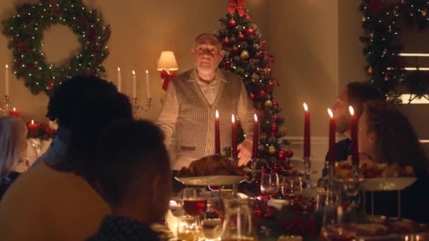 老爷爷给他的多文化大家庭讲笑话或生活故事 他们庆祝圣诞节 有盘子和蜡烛的餐桌 家中温馨的圣诞大餐气氛 — 图库视频影像