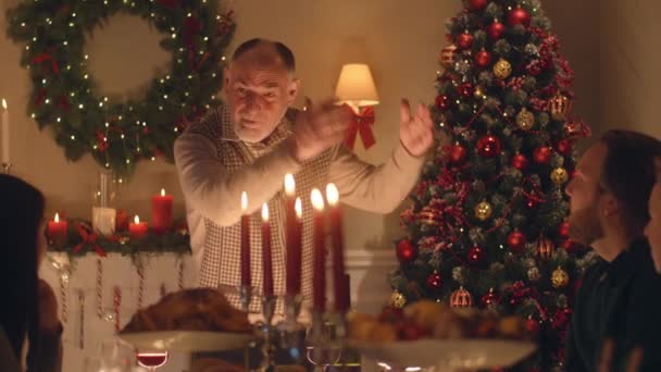 老人は大規模な多文化家族に冗談や人生の話をします クリスマスを祝うんだ 食器やキャンドル付きのテーブルを提供します 家庭でのクリスマスディナーの暖かい雰囲気 — ストック動画