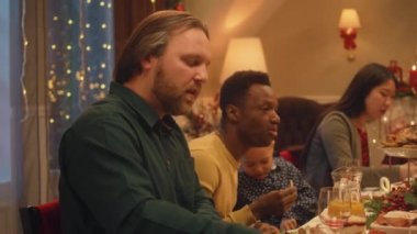 Mutlu çok kültürlü aileler Noel 'i ya da 2023 yılını kutluyor, konuşuyor ve yiyor. Farklı yemek ve mumlarla servis edilen tatil masası. Aile içinde sıcak bir Noel yemeği..