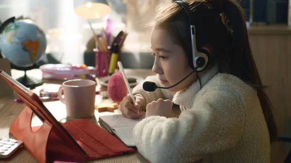 stock image Young asian girl sits at the table in headphones, talks on online conference. Schoolgirl listens teacher, mark new information, writes homework. Distance learning concept. Remote studying.