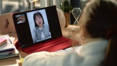 Genç kız evinde rahat bir masada oturuyor ve yetişkin Asyalı öğretmenle konuşuyor. Öğrenci çevrimiçi ders dinler, yeni materyaller yazar ve tablete çalışmalar yapar. Uzaktan konferans. Arka plan.