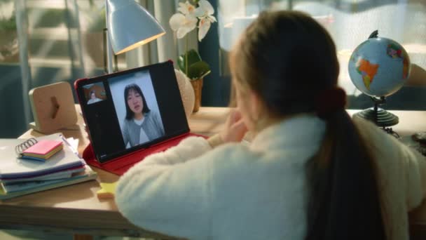 年轻姑娘舒适地坐在餐桌旁 与亚洲的成年教师交谈 学生在网上听课 写下新材料 并在平板电脑上学习 远程会议 背景色 — 图库视频影像