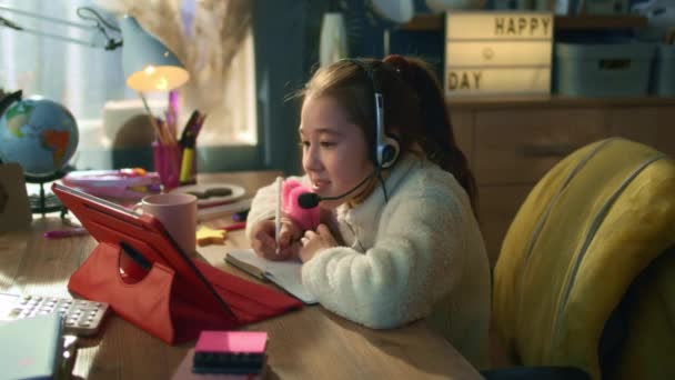 若いアジアの女の子は ヘッドフォンでテーブルに座って オンライン会議で話をします 女子高生は授業の終わりに先生に拍手を送り 宿題を書きます 距離学習の概念 遠隔研究 — ストック動画