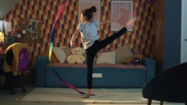 Genç Asyalı kız jimnastik kurdelesiyle dans ediyor, dikey bacak ayrımını gösteriyor ve kendi etrafında dönüyor. Jimnastik eğitimi. Spor hobisi. Karantinadaki fiziksel sağlığı yükseltiyorum. Yavaş çekim.