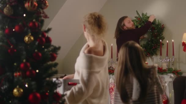 ボールでクリスマスツリーを話して飾るお母さんと娘 父は壁にクリスマスの花輪を掛けます 冬の休暇のために家を準備する家族 幸せな若い女の子 スローモーション — ストック動画