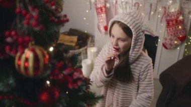 Hafif pijamalı genç bir kız Noel ağacına gelir, şeker yalar ve topları ve oyuncakları düzeltir. Duvarda çelenkler parıldıyor. Kış tatilinden önce hazırlanıyorum. Yeni yılın büyüsü. Yavaş çekim.