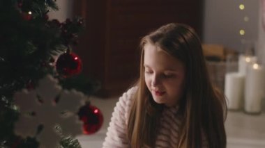 Hafif pijamalı genç bir kız yere oturuyor, Noel ağacını toplar ve oyuncaklarla süslüyor, gülümsüyor ve boş vakitlerini evde geçiriyor. Çocuk kış tatili ve yeni yıldan önce odayı hazırlar. Yavaş çekim.