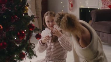 Hafif pijamalı genç kız Noel ağacını dallarla süslüyor. Anne, kız kulağına kar tanesi oyuncağı giydir. Aile Noel tatilinden önce eğleniyor ve oturma odasını hazırlıyor. Yavaş çekim