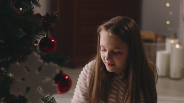 穿着淡淡的睡衣的小女孩坐在地板上 用球和玩具装饰圣诞树 微笑着待在家里 孩子们在寒假和新年前准备好房间 慢动作 — 图库视频影像