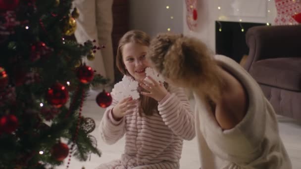年轻姑娘穿着淡淡的睡衣 用树枝装饰圣诞树 妈妈把雪花玩具放在女儿的耳朵上 全家在圣诞假期前玩乐和准备客厅 慢动作 — 图库视频影像