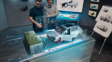 İki Mühendis Geliştirici, geleceğin holografik masasının yanındaki tasarım stüdyosunda duruyorlar ve 3 boyutlu bir araba kazası test simülatöründe test yapıyorlar.
