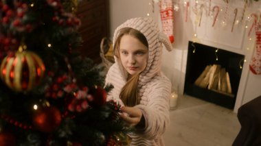 Hafif pijamalı genç bir kız Noel ağacına gelir, şeker yalar ve topları ve oyuncakları düzeltir. Duvarda çelenkler parıldıyor. Kış tatilinden önce hazırlanıyorum. Yeni yılın büyüsü. Yavaş çekim.