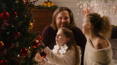 Aile birlikte oturma odasında oturuyor ve Noel ağacını dallarla süslüyor. Duvarda yanıp sönen çelenk ışıkları. Kışın sıcacık bir evde tatil. Yavaş çekim