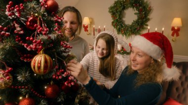 Young girl with parents choosing toys and decorating Christmas tree. Family preparing living room before winter holidays or New Year. Atmosphere of Christmas at cozy home. Slow motion. clipart