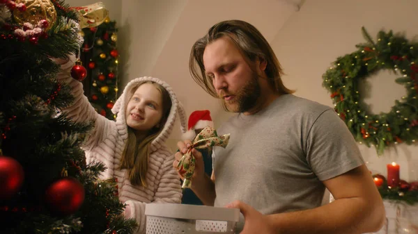 stock image Daughter with dad choosing balls and toys, decorating Christmas tree. Young girl in pajama helps father with preparing house before Christmas holidays. Magic of winter at cozy home. Slow motion.