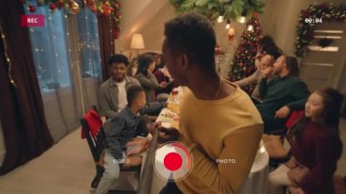 Özçekim çubuğu videosu olan Afro-Amerikalı bir adam, tabaklarla dolu bir masa gösteriyor. Noel 'i ya da yeni yılı kutlayan çok kültürlü bir aile. Evde aile Noel yemeği atmosferi. Kamera görünümü.